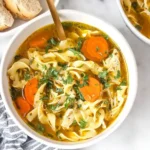 Bowl of slow cooker chicken noodle soup with carrots, egg noodles, and fresh herbs, served with bread on the side.