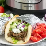Slow Cooker Beef Gyros wrapped in foil with shredded beef, fresh cucumber, tomato, red onion, lettuce, and tzatziki sauce on a plate.