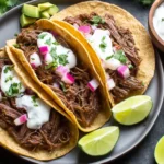 Juicy chuck roast tacos with shredded beef, sour cream, onions, and lime wedges on a plate.