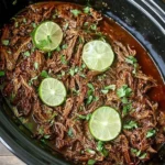 Easy Slow Cooker Barbacoa with shredded beef, garnished with lime slices and fresh cilantro in a black slow cooker.