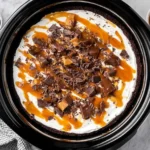 Top view of Slow Cooker Better Than Sex Cake topped with Cool Whip, caramel drizzle, chopped Heath bars, and chocolate sauce in a black slow cooker.