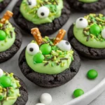 Spooky Cauldron Cookies topped with green frosting, candy eyeballs, sprinkles, and pretzel stir sticks on a white plate.