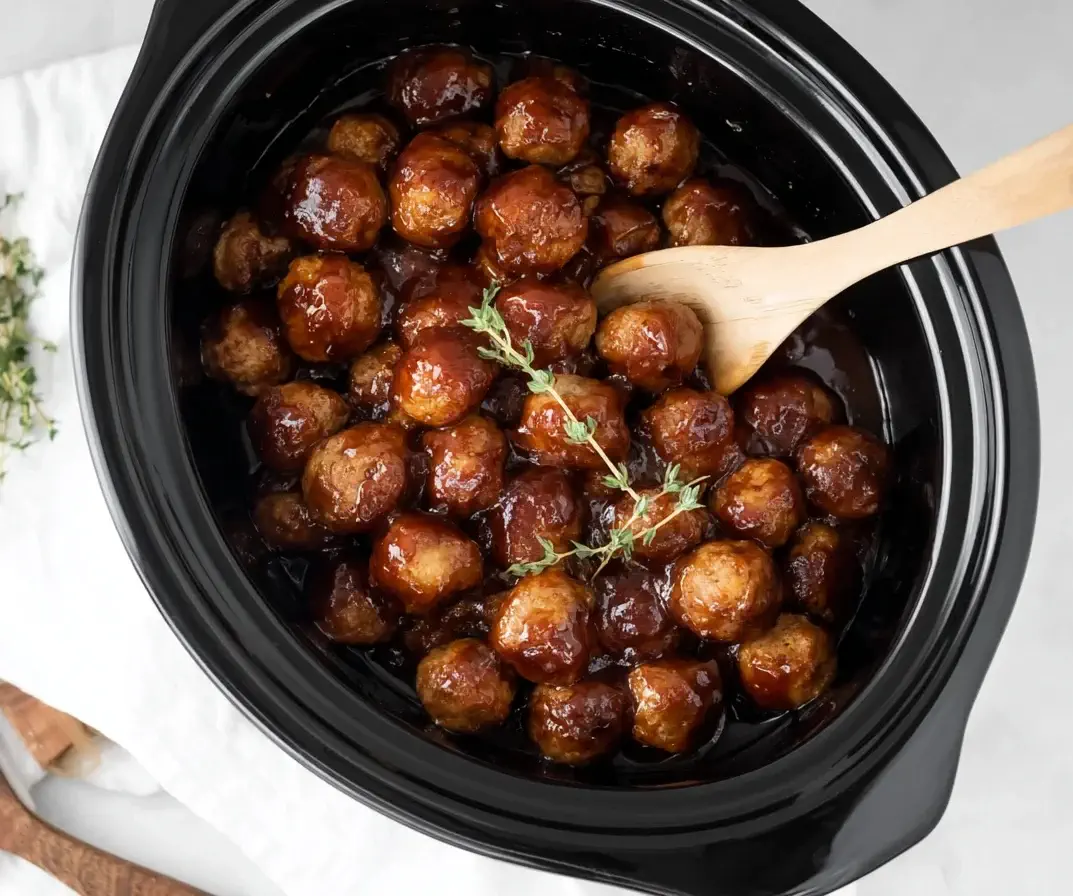 Slow Cooker Apple Butter Meatballs coated in a sweet and tangy apple butter sauce inside a black crockpot with a wooden spoon.