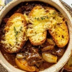 Bowl of French Onion Beef Stew topped with golden baguette slices, melted Gruyere cheese, and fresh thyme on a rustic table setting.