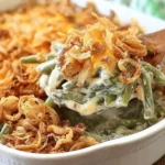 Creamy Green Bean Casserole being served with a wooden spoon, topped with melted cheese and crispy fried onions in a white baking dish.