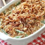 Homemade Green Bean Casserole topped with crispy fried onions in a white baking dish on a red checkered cloth.
