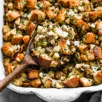 Homemade Thanksgiving stuffing with golden bread cubes, herbs, and sausage served in a rustic white baking dish.