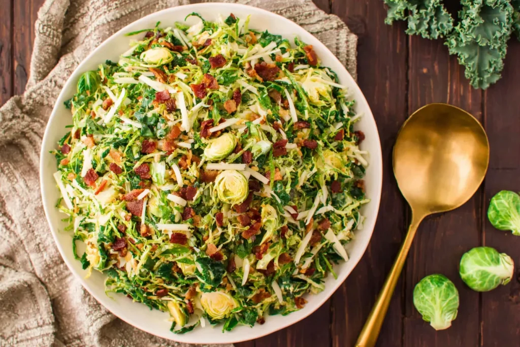 Overhead view of Brussels Sprout, Kale + Apple Salad with Sharp Cheddar + Bacon in a white bowl, topped with apples, bacon, and cheese.
