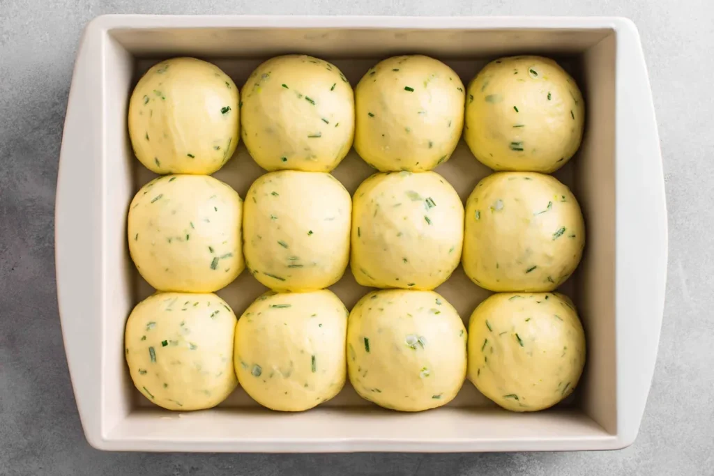 Garlic Bread Rolls dough arranged in a 9x13 baking pan before rising, speckled with fresh herbs and ready to proof.