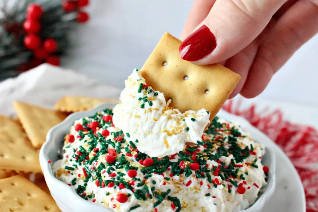 Hand dipping a cracker into creamy Little Debbie Christmas Tree Cake Dip topped with festive holiday sprinkles.