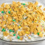 No Bake Christmas Cookie Lush topped with whipped cream, crushed cookies, sprinkles, and holiday candies in a glass baking dish.