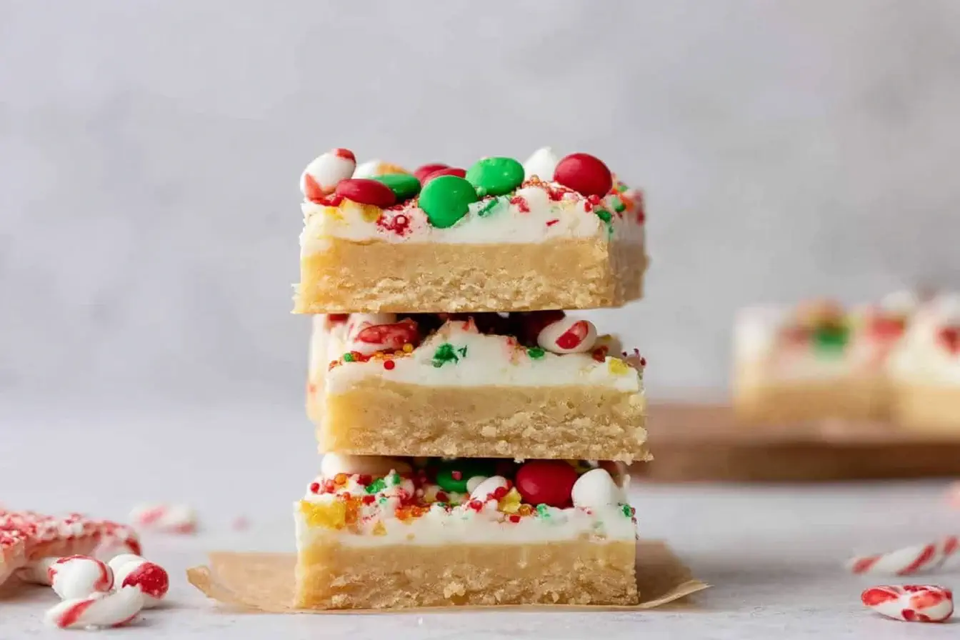 Stack of festive Christmas Sugar Cookie Bars topped with white frosting, holiday sprinkles, and candies on a light background.