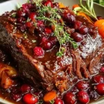Slow-Braised Beef Roast with Cranberry Balsamic Glaze topped with fresh herbs, served over mashed potatoes with carrots and rich sauce.