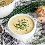Slow Cooker Potato Leek Soup in a white soup tureen topped with chives and served with fresh bread and herbs.
