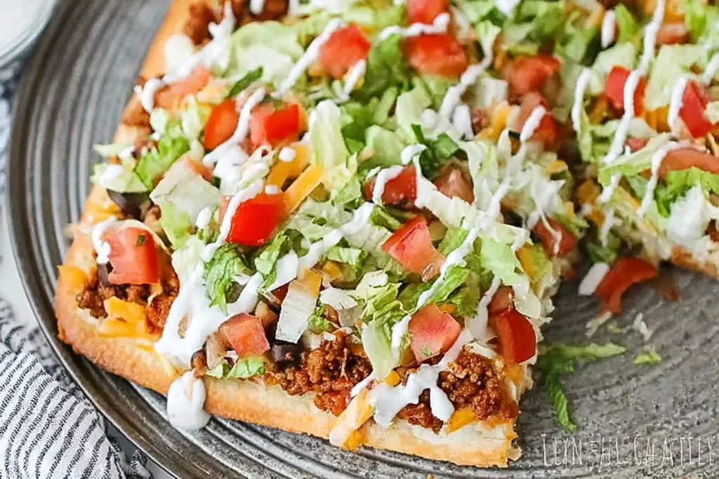 Taco Pizza close-up slice topped with taco-seasoned ground beef, shredded lettuce, diced tomatoes, melted Mexican cheese, and a sour cream drizzle on a plate.