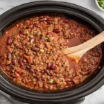 Chili Crockpot recipe cooking in a slow cooker with ground beef, beans, and rich tomato sauce being stirred with a wooden spoon.