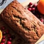 Freshly baked Christmas Bread loaf surrounded by cranberries and oranges on a wooden board, showcasing its golden crust and festive flavors.