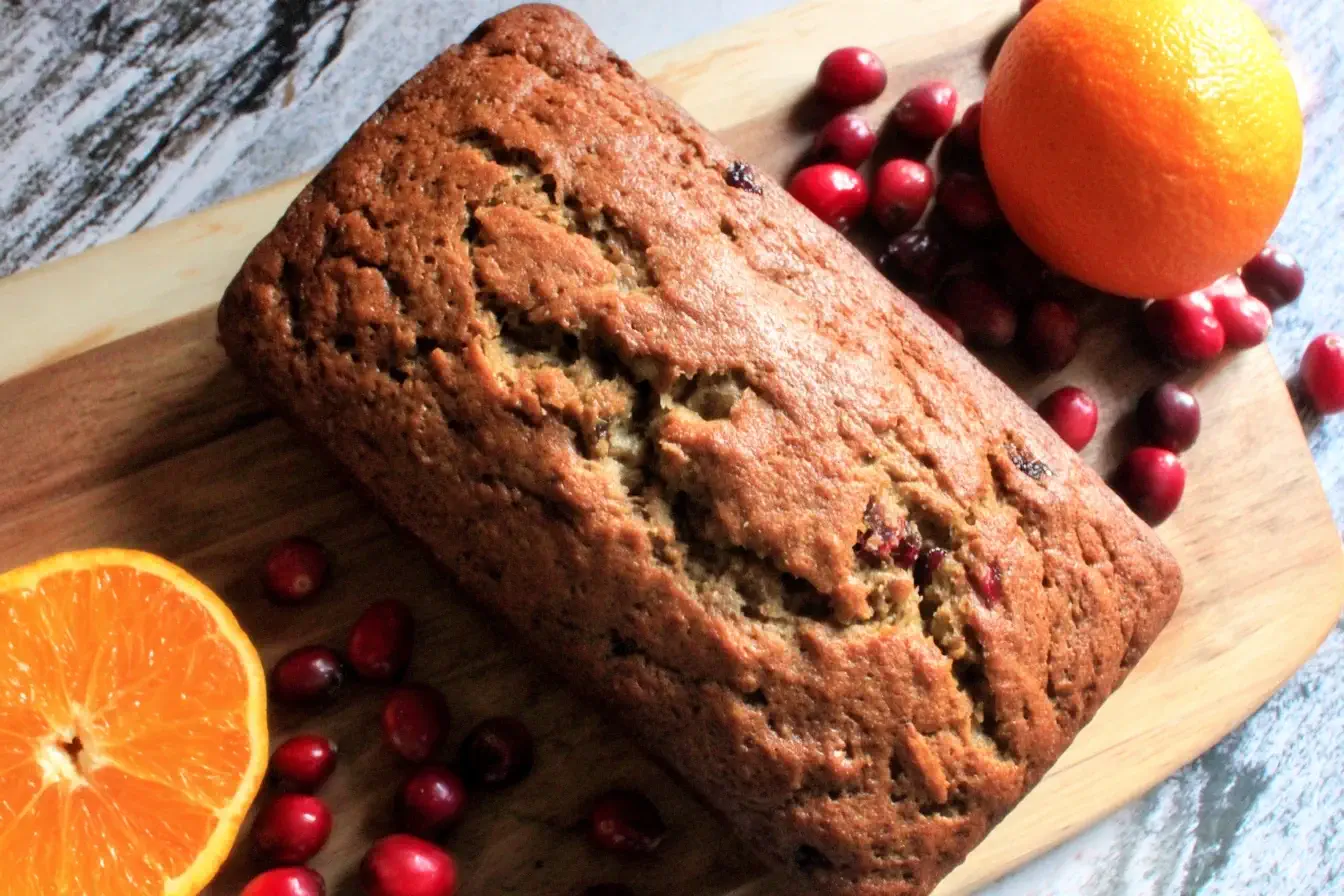 Freshly baked Christmas Bread loaf surrounded by cranberries and oranges on a wooden board, showcasing its golden crust and festive flavors.