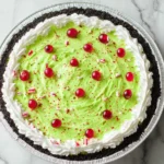 Top view of a No Bake Grinch Pie with bright green filling, whipped cream border, and festive red candies in an Oreo crust.