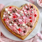 Heart Shaped Cookie Cake topped with pink frosting, candy hearts, and chocolate pieces for Valentine’s Day