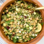 The Jennifer Aniston Salad in a wooden bowl with quinoa, chickpeas, cucumber, fresh herbs, feta, and lemon wedge.