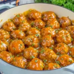 Awesome Sauce Party Meatballs simmering in a Dutch oven coated in glossy sweet and spicy sauce with fresh parsley garnish