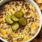 Crockpot Big Mac Dip in a white bowl topped with sliced pickles, with melted cheese and seasoned ground beef, surrounded by tortilla chips