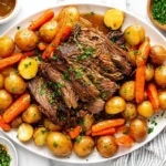 Easy Crock Pot Pot Roast on a platter with tender chuck roast, baby potatoes, carrots, and savory gravy, finished with fresh parsley.