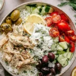 Slow Cooker Greek Chicken Recipe bowl with shredded lemon herb chicken, rice, tzatziki, cucumbers, tomatoes, and olives.