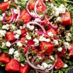 Feta Watermelon Salad on arugula with crumbled feta, red onion, fresh herbs, and balsamic glaze in a black bowl.