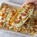 Hand holding Baked Chicken Bacon Ranch Tacos topped with lettuce, melted cheese, crispy bacon, and ranch drizzle in a baking dish.