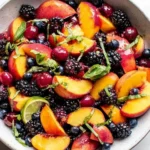 Honey Lime Basil Peach Fruit Salad with juicy sliced peaches, blackberries, blueberries, pomegranate seeds, fresh basil ribbons, and a lime wedge in a bowl.