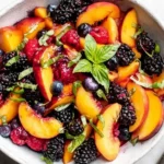 Honey Lime Basil Peach Fruit Salad in a gray bowl with sliced peaches, blackberries, blueberries, raspberries, and fresh basil leaves on top.