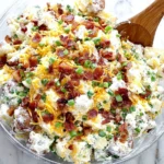 Loaded Baked Potato Salad in a clear bowl, topped with crispy bacon, shredded cheddar, and green onions, with a wooden spoon on a marble counter.
