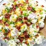 Loaded Baked Potato Salad in a glass bowl topped with crispy bacon, shredded cheddar, and chopped green onions on a light marble background.