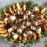 Peach and Burrata Salad with arugula, sliced peaches, cherries, burrata cheese, chopped nuts, fresh herbs, and balsamic glaze on a large platter.