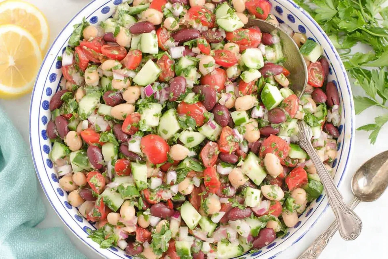 Mediterranean Bean Salad in a white bowl with chickpeas, kidney beans, cucumbers, tomatoes, herbs, and lemon slices.