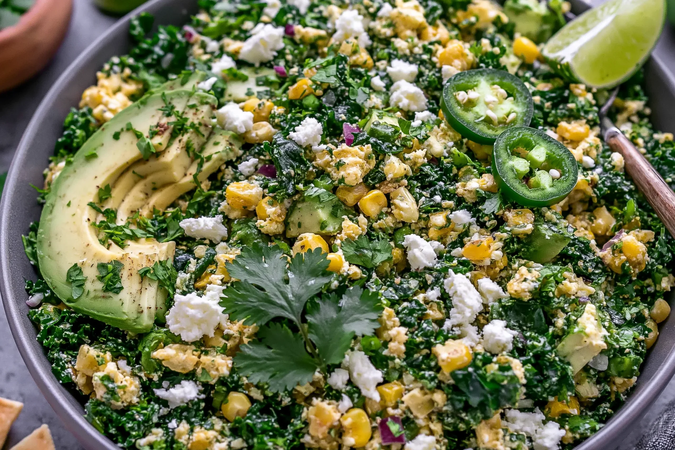 Mexican Street Corn Kale Salad with avocado, cotija cheese, corn, cilantro, jalapeño slices, and lime in a bowl