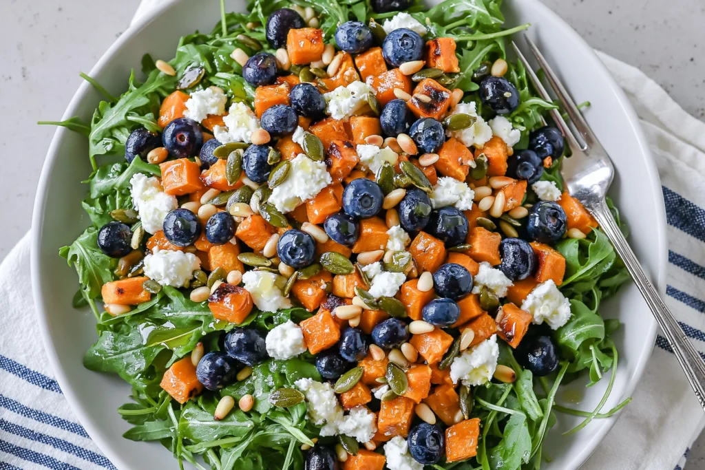 Honey Sweet Potato Summer Salad in a white bowl with arugula, roasted sweet potato cubes, blueberries, goat cheese, pepitas, pine nuts, and honey lemon vinaigrette.