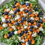 Honey Sweet Potato Summer Salad in a white bowl with arugula, roasted sweet potato cubes, blueberries, goat cheese, pepitas, pine nuts, and honey lemon vinaigrette.