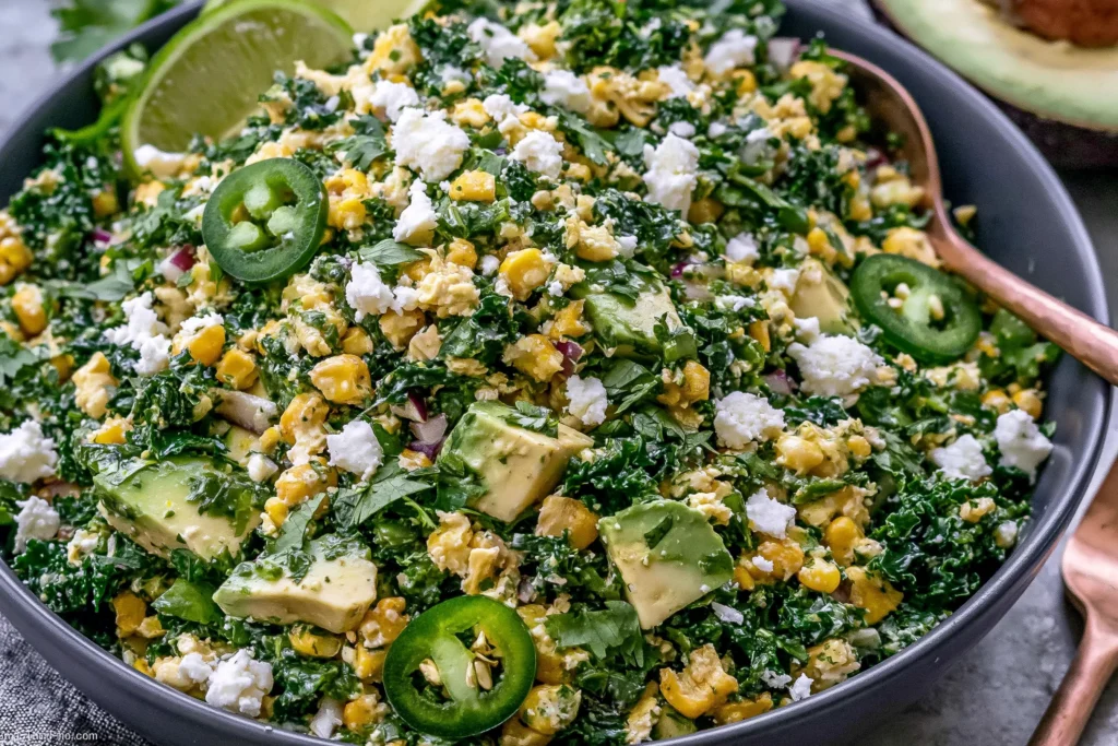 Mexican Street Corn Kale Salad with corn, avocado chunks, cotija cheese, cilantro, jalapeño slices, and lime in a dark bowl