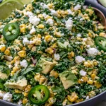 Mexican Street Corn Kale Salad with corn, avocado chunks, cotija cheese, cilantro, jalapeño slices, and lime in a dark bowl